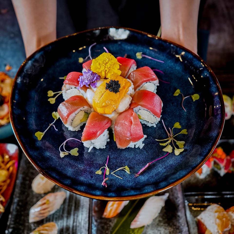 Hands holding a dark blue plate of Sushiya's tuna sushi rolls, uni, and black caviar, garnished with flowers.