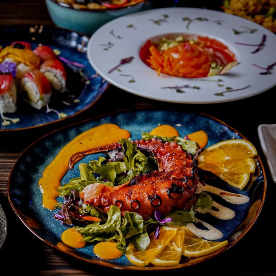 Grilled octopus with salad and orange slices on a blue plate, with sushi and another dish in the background.