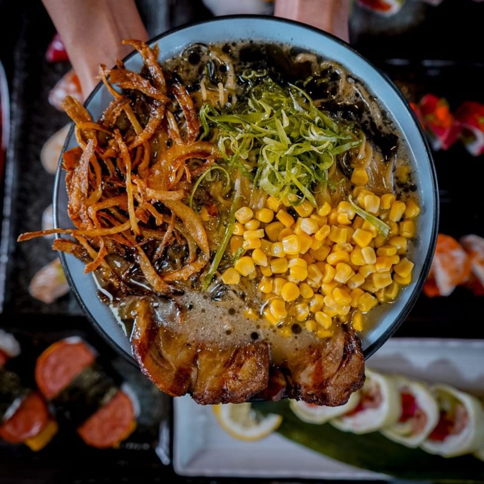 Hands holding a bowl of ramen with pork, corn, scallions, and crispy onions.